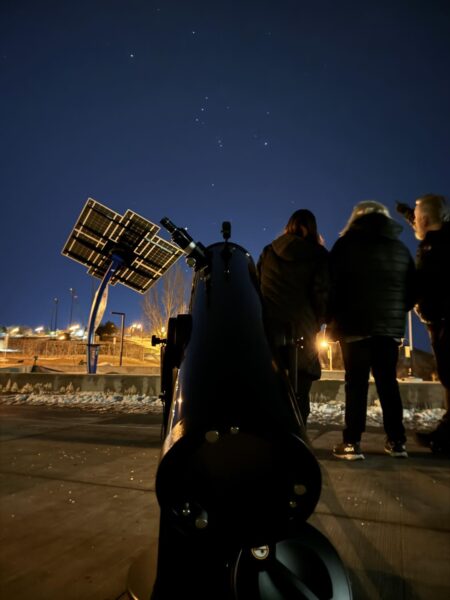 Photo with telescope in the foreground and 3 adults standing in the background looking up at the night sky.