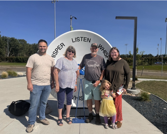 Grand parents, parents, and grandchild standing together in front of whisper dishes outside of the science center on the walkway.