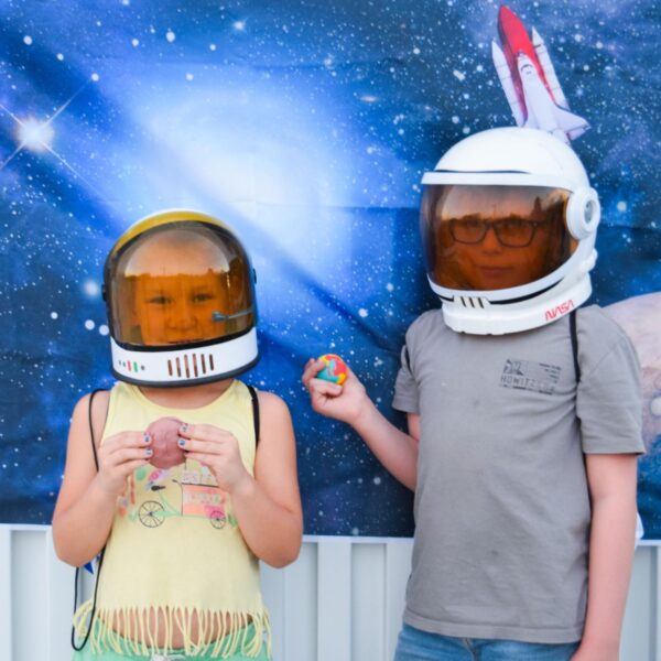 Two kids with astronaut helmets on in front of a space backdrop.